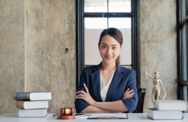 smiling Asian woman in a white shirt and blue blazer with arms crossed sitting at a table with books and small lady justice statue