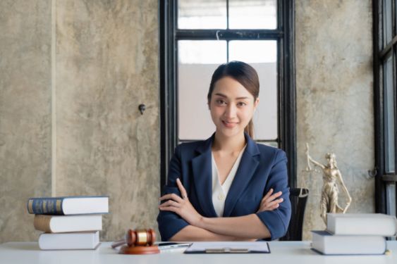 smiling Asian woman in a white shirt and blue blazer with arms crossed sitting at a table with books and small lady justice statue